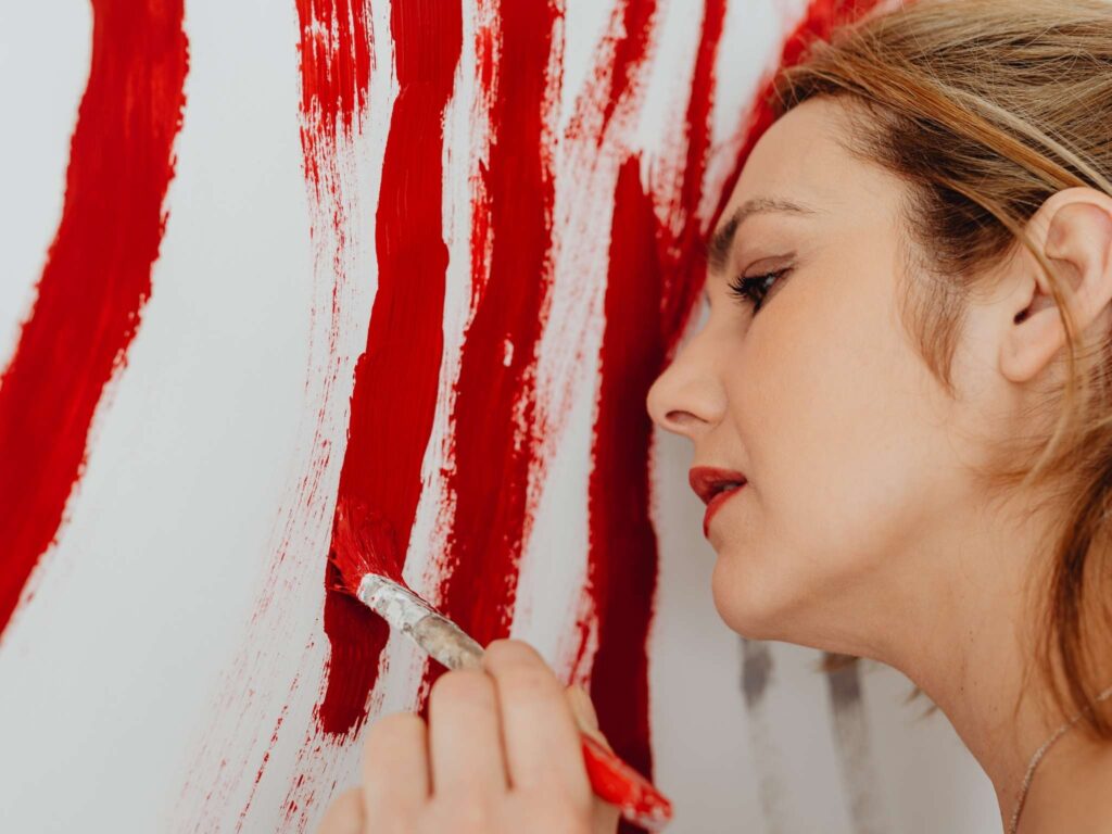 Eine Frau trägt mit einem Pinsel rote Farbe auf eine vertikale Fläche auf.