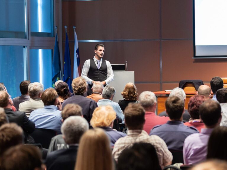 Eine große Gruppe von Teilnehmenden sitzt im Saal, während ein Mann vorne einen Vortrag hält.
