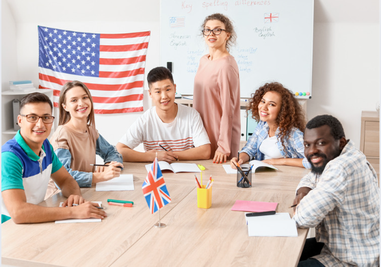 Gruppe internationaler Lernender in einem Sprachkurs sitzt lächelnd am Tisch mit Notizbüchern und Flaggen, während die Lehrkraft Motivation und Sprachpraxis fördert.