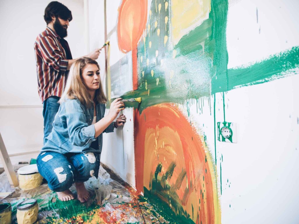 Eine Frau und ein Mann malen mit Pinseln auf eine Wand, umgeben von vielen Farbdosen.