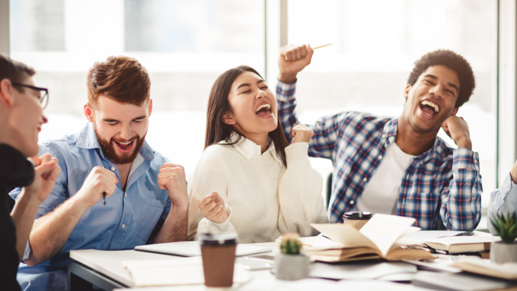 Eine Gruppe junger Erwachsener sitzt gemeinsam am Tisch und jubelt begeistert. Alle wirken fröhlich und motiviert, einige heben die Arme, andere lachen laut. Die Szene vermittelt Energie, Teamgeist und Erfolgserlebnisse in einer Lern- oder Arbeitssituation.