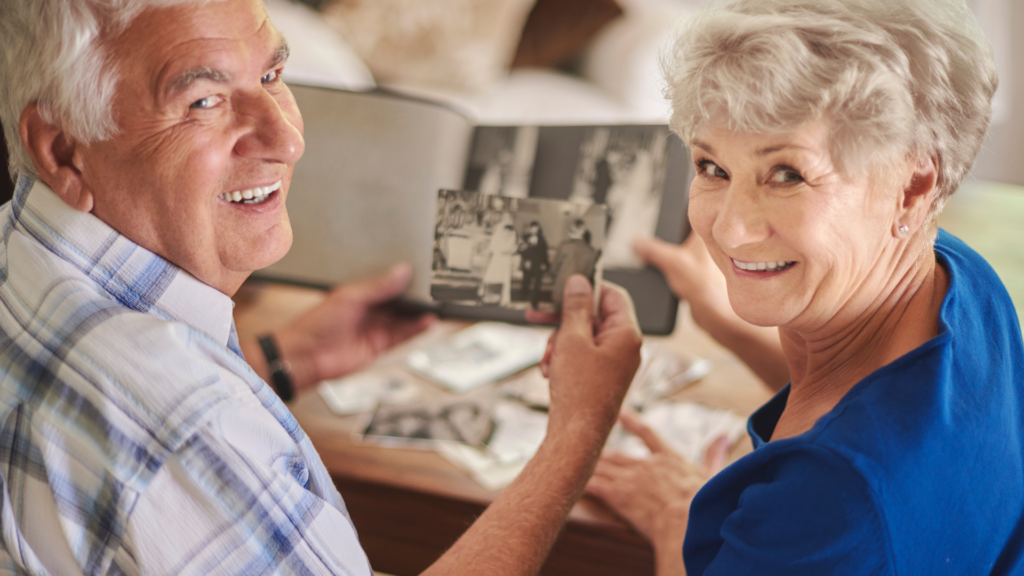 Älteres Paar sitzt gemeinsam am Tisch, betrachtet alte Fotos in einem Album und lächelt – sie wirken zufrieden, verbunden und voller schöner Erinnerungen.