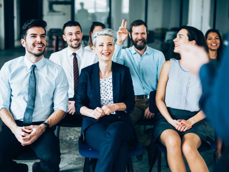 Gruppe von Menschen sitzt in einem Büro, lächelt und eine Person meldet sich mit erhobener Hand.