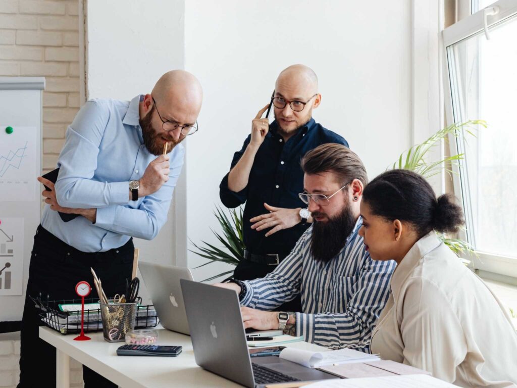 Mehrere Personen unterschiedlicher Kulturen arbeiten gemeinsam an einem Schreibtisch, eine Person telefoniert, andere schauen auf Laptops.