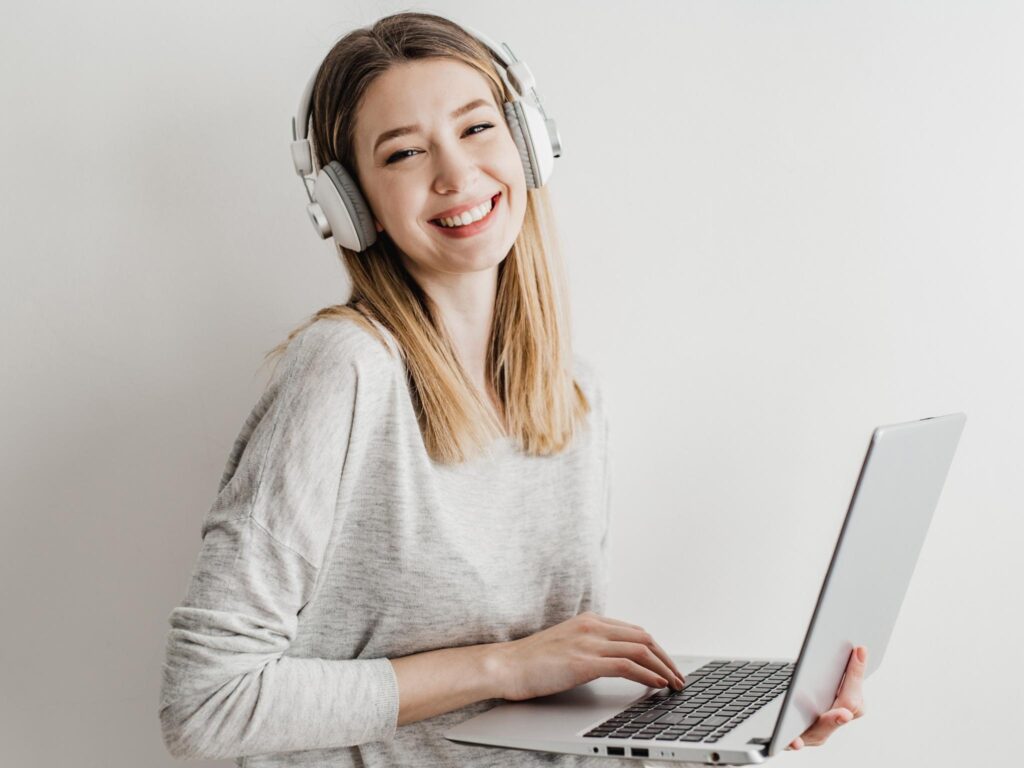 Junge Frau mit Kopfhörern hält einen Laptop und lächelt.