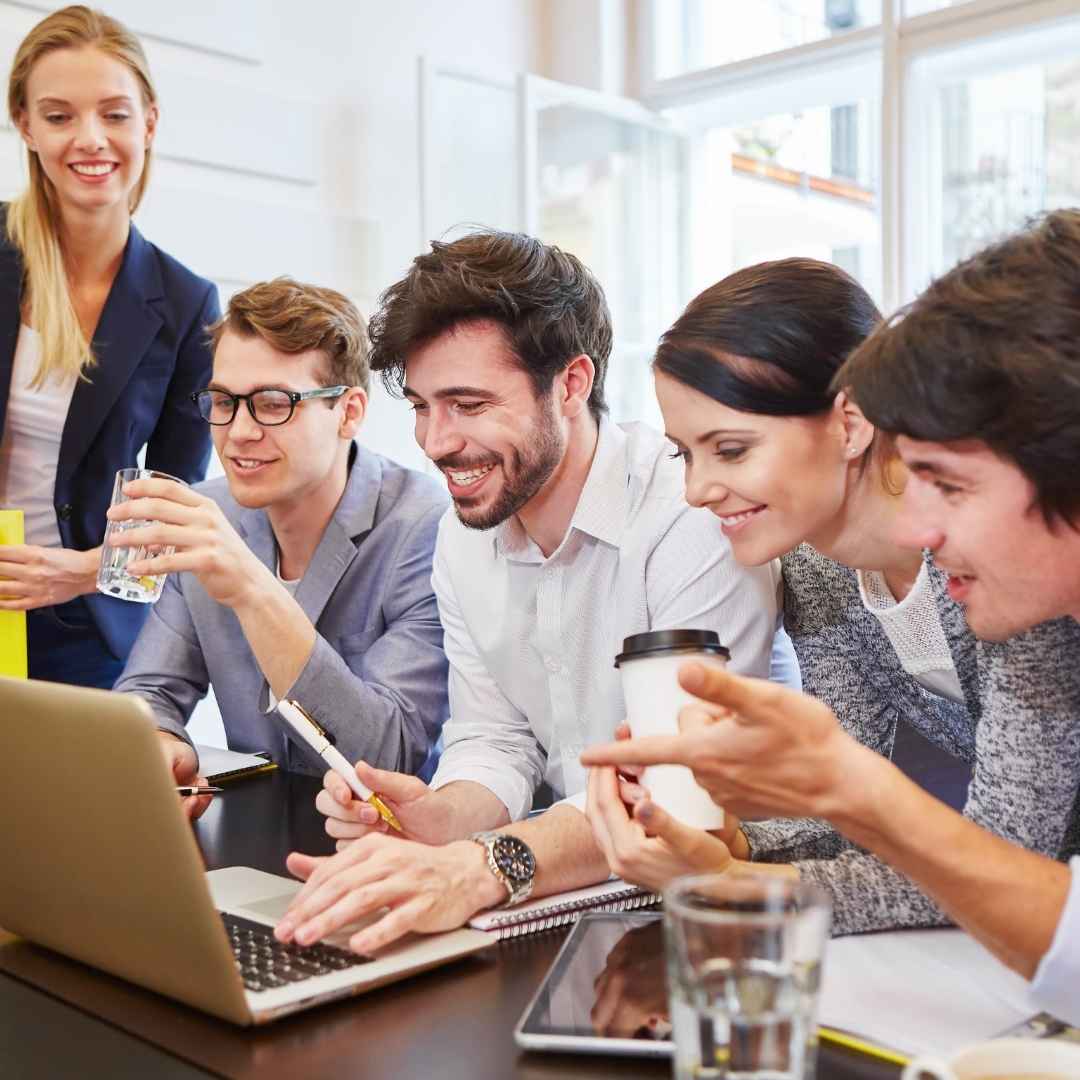 Eine Gruppe junger Erwachsener sitzt gemeinsam um einen Laptop und arbeitet konzentriert an einem Text. Alle wirken engagiert, lachen und tauschen sich aus. Vor ihnen liegen Notizbücher, Stifte und Getränke. Eine Frau steht daneben und beobachtet die Runde lächelnd. Die Atmosphäre ist offen, kollaborativ und kreativ.