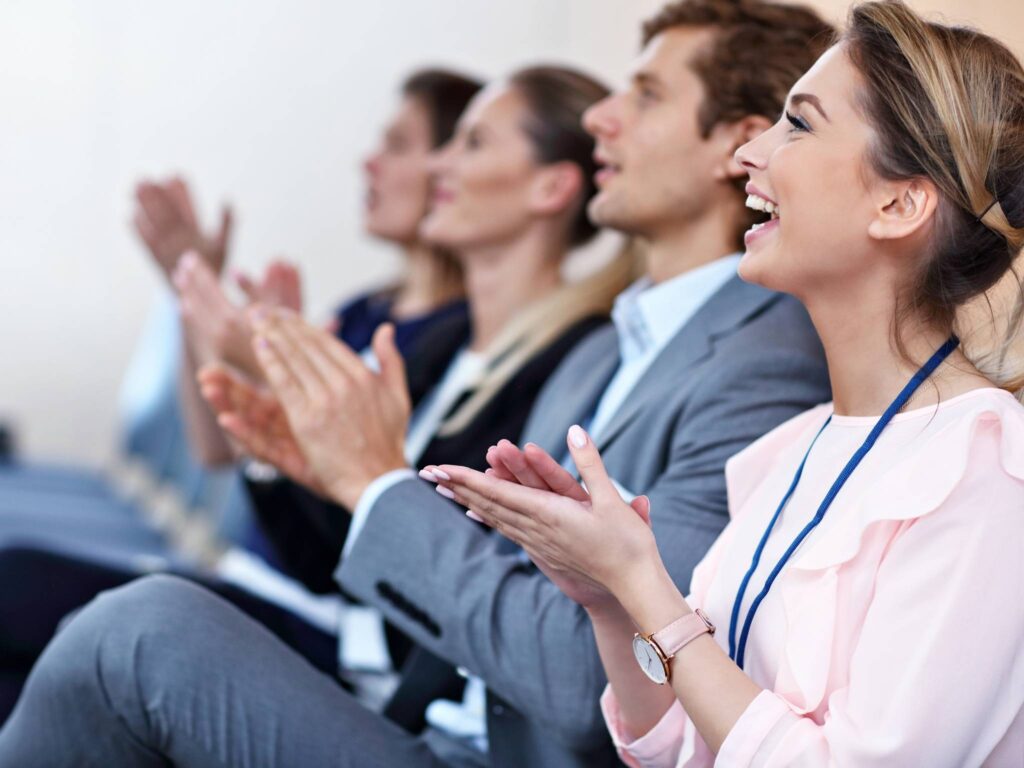 Mehrere Menschen sitzen bei einer Konferenz, lächeln und klatschen, vorne lacht eine Frau besonders sichtbar.