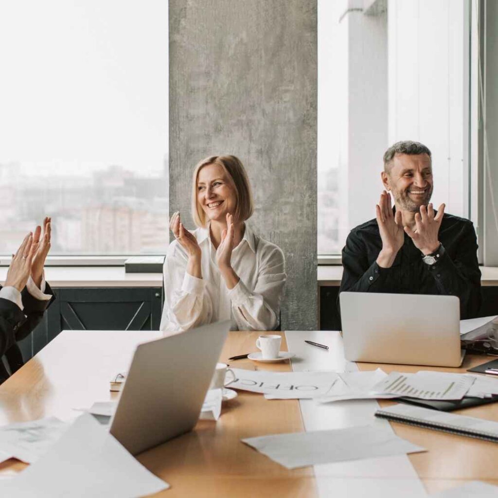 Mehrere Erwachsene sitzen an einem Tisch in einem hellen Seminarraum. Sie klatschen und lächeln, auf dem Tisch liegen Laptops, Notizen und Unterlagen. Die Szene zeigt Wertschätzung, Austausch und positive Gruppendynamik in der Erwachsenenbildung.