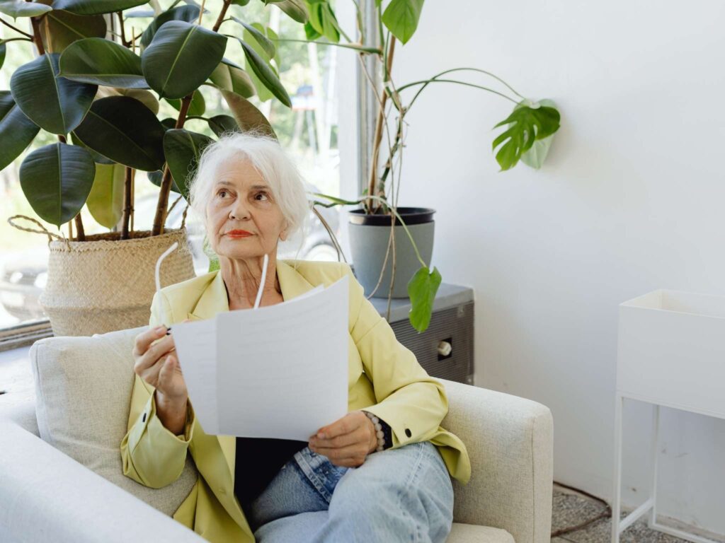 Seniorin sitzt im Sessel, hält Karten und Brille, denkt nach und arbeitet aktiv mit KI-Szenarien.