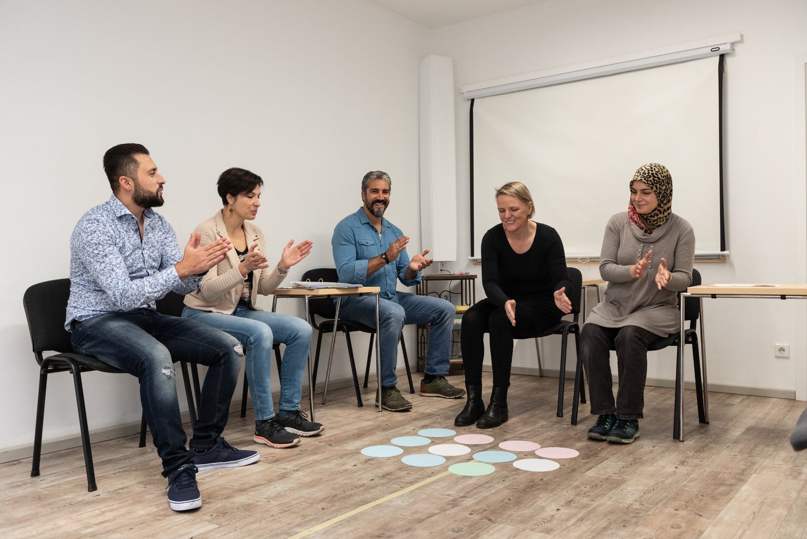 Gruppe im Stuhlkreis klatscht, auf dem Boden liegen bunte Kreise